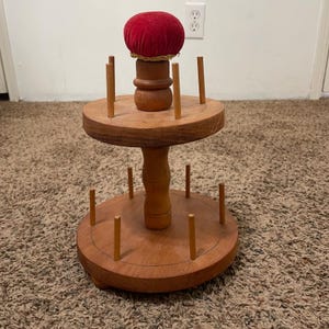 Könnte beinhalten: Ein hölzerner Garnrollenhalter mit einem roten Samt-Nadelkissen oben. Der Garnrollenhalter hat zwei kreisförmige Plattformen mit Holzstiften zum Halten von Garnrollen. Das Nadelkissen ist rund und hat eine goldene Umrandung.