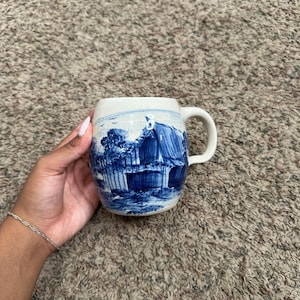Puede incluir: Una taza de cerámica con base de color blanco roto moteado y una escena de paisaje pintada a mano en azul que muestra una casa y árboles. La taza tiene un cuerpo redondeado, un asa curva lisa y un acabado brillante con una ilustración azul.