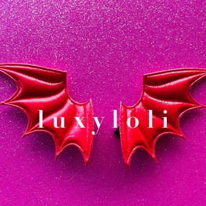 Puede incluir: Un par de alas de diablo rojas brillantes, con una superficie texturizada, sobre un fondo púrpura brillante. Las alas son simétricas y detalladas, con puntas puntiagudas. El texto "luxyloli" está en blanco.