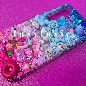 Peut inclure: Une coque de téléphone rose, bleue et blanche décorée de divers breloques, dont un donut, un flamant rose, un pingouin et une licorne. La coque est recouverte de strass et de paillettes.