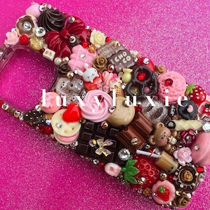 Könnte beinhalten: Eine rosa und weiße Handyhülle, die mit einer Vielzahl von Miniatur-Lebensmitteln verziert ist, darunter Schokoladentafeln, Kekse, Erdbeeren und ein rosa und weißer Donut. Die Hülle ist mit Strasssteinen besetzt und hat eine transparente Rückseite.