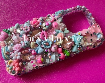 May include: A pink and blue phone case decorated with rhinestones, bows, flowers, and other cute charms. The case has a clear back and a cutout for the camera.