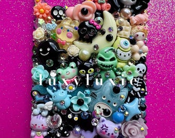 May include: A decorative phone case with a multitude of colorful, whimsical charms. The case is adorned with various figures, including skulls, cats, and flowers, and is embellished with rhinestones. The text "luxy luxie" is visible.