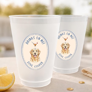 May include: Two translucent frosted plastic cups featuring a circular graphic of a golden retriever with a cocktail glass balanced on its head. The text reads "DRINKS ON ME!" and "LOVE, COOPER" in blue lettering within a light blue oval border.