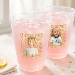 May include: Two clear plastic cups filled with pink liquid and ice. Each cup has a gold "CLASS OF 2026" design with a child's photo. One cup has the name "Jason" and the other has the text "WE ARE THE FUTURE".
