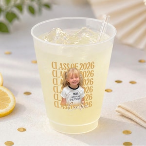 May include: A clear plastic cup filled with a yellow beverage, ice cubes, and a straw. The cup has a gold design that says "CLASS OF 2026" and a photo of a young girl wearing a shirt that says "WE ARE THE FUTURE". Sliced lemons and gold confetti are also visible.