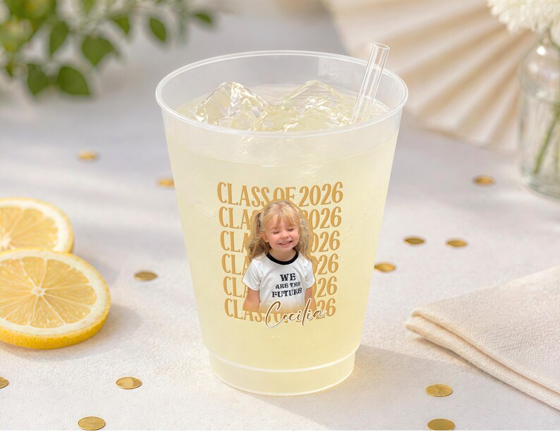 May include: A clear plastic cup filled with a yellow beverage, ice cubes, and a straw. The cup has a gold design that says "CLASS OF 2026" and a photo of a young girl wearing a shirt that says "WE ARE THE FUTURE". Sliced lemons and gold confetti are also visible.