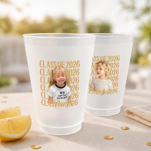 May include: Two white plastic cups with gold text that reads "CLASS OF 2026". Each cup has a photo of a child. One cup has the text "WE ARE THE FUTURE". Slices of lemon and gold confetti are in the foreground.