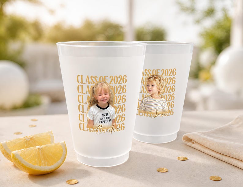 May include: Two white plastic cups with gold text that reads "CLASS OF 2026". Each cup has a photo of a child. One cup has the text "WE ARE THE FUTURE". Slices of lemon and gold confetti are in the foreground.