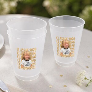 May include: Two frosted plastic cups with a gold "CLASS OF 2026" design and a photo of a young girl. The cups are on a white surface with a fork, knife, and plate in the background. A small flower arrangement is also visible.