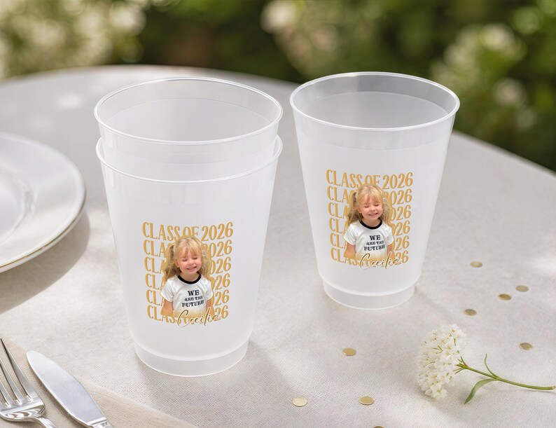 May include: Two frosted plastic cups with a gold "CLASS OF 2026" design and a photo of a young girl. The cups are on a white surface with a fork, knife, and plate in the background. A small flower arrangement is also visible.