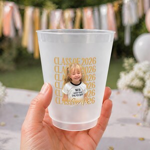 May include: A frosted plastic cup with a photo of a smiling child wearing a shirt that says "We are the future". The cup has "Class of 2026" printed in gold, along with the child's name. The cup is held in a hand.