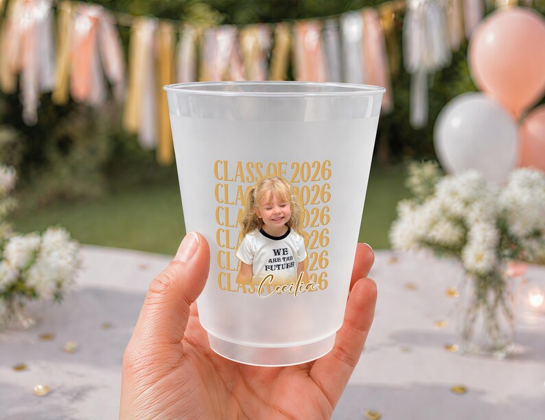 May include: A frosted plastic cup with a photo of a smiling child wearing a shirt that says "We are the future". The cup has "Class of 2026" printed in gold, along with the child's name. The cup is held in a hand.