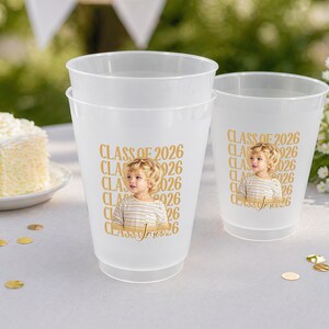 May include: Three frosted plastic cups with a gold design. The design includes the text "CLASS OF 2026" and a photo of a child. The cups are on a white surface with gold confetti and a piece of cake.