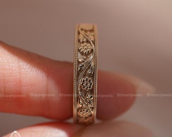 Puede incluir: Un anillo de banda de oro con un intrincado patrón floral y de enredaderas en relieve en la superficie exterior, enmarcado por delicados bordes de cuentas. El metal tiene un acabado pulido y cálido con motivos florales.