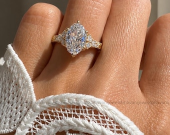 Pode incluir: Um anel de ouro com um grande diamante central de corte marquise em cravação de garras, ladeado por diamantes redondos menores em cada lado da banda. A banda tem um acabamento em ouro polido e o anel é usado num dedo.