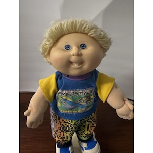 May include: A doll with blonde yarn hair, blue eyes, and a single visible tooth. It wears a blue shirt with yellow sleeves featuring a graphic print that says FREESTYLE LPK, paired with patterned pants in black, yellow, and orange, and blue sneakers.
