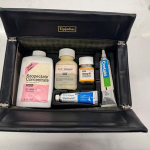 May include: A black leatherette case with the Upjohn logo, open to reveal several medicine bottles and tubes. The bottles include Kaopectate Concentrate, P-A-C Compound, and Unicap M. A tube of Cortaid and Myciacin are also visible.