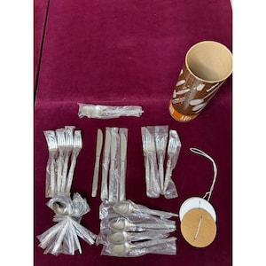 May include: A collection of silver-toned stainless steel flatware including forks, knives, and spoons, most individually wrapped in clear plastic. A cylindrical cardboard storage tube and two circular cardboard discs with white string are included.