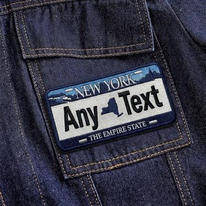 May include: Close-up of a dark blue denim jacket with a rectangular patch. The patch features the text "NEW YORK" above "Any Text" and "THE EMPIRE STATE" with a silhouette of New York state.