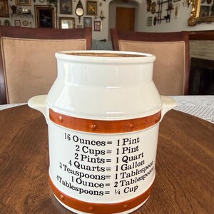 May include: A cream-colored ceramic canister shaped like a milk can with two side handles. It features two horizontal orange bands with raised dots and black printed text listing kitchen measurement conversions including 16 Ounces equals 1 Pint.