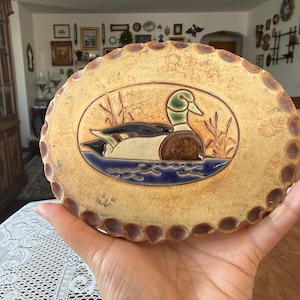 May include: An oval ceramic wall plaque with a scalloped edge, featuring a relief illustration of a mallard duck swimming in blue water among reeds. The piece has a tan, speckled finish with a signature visible on the lower left side.