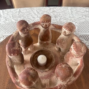 Op de afbeelding: Een ronde terracotta kaarsenhouder met zes gestileerde figuren die in een cirkel staan met hun armen in elkaar geslagen rond een centrale kom. De figuren hebben een roodbruine afwerking met lichtere, getextureerde oppervlakken. Een klein wit waxinelichtje staat in het midden.