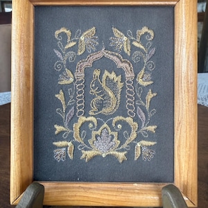 May include: A framed embroidery piece featuring a central squirrel motif enclosed within an arched structure, surrounded by intricate gold and silver floral patterns on a dark fabric background, all encased in a light-toned wooden frame.