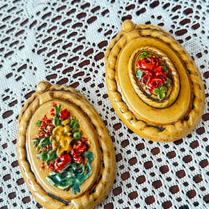 May include: Two oval-shaped ceramic wall plaques with a mustard yellow finish and a raised, hand-painted floral design. One plaque features a central bouquet of red and yellow flowers, while the other displays a cluster of red roses.