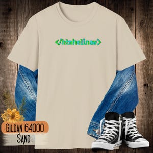 Op de afbeelding: Een zandkleurig t-shirt met korte mouwen en de tekst htmhellnaw tussen groene en blauwe code-haakjes op de borst. Het shirt is gelaagd over een blauw spijkerjack en gecombineerd met zwart-witte hoge canvas sneakers.