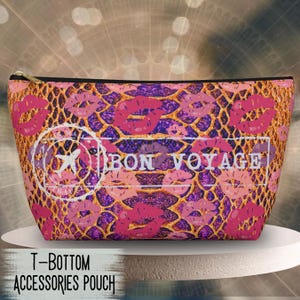May include: A zippered accessories pouch featuring a purple and gold snakeskin pattern overlaid with pink lip prints. A white rectangular graphic on the front displays the text BON VOYAGE alongside a circular stamp icon containing an airplane.
