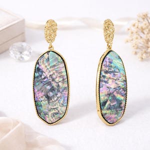 May include: A pair of gold-toned earrings with iridescent oval-shaped pendants. The pendants have a shimmering, multi-colored surface, framed by a gold rope-like border. The top of each earring features a textured, ornate gold design.