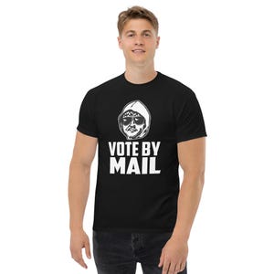 May include: Black t-shirt with a white graphic of a person in a hooded sweatshirt and sunglasses. The text "VOTE BY MAIL" is printed in bold, white letters below the graphic. The shirt is a classic crew neck style.