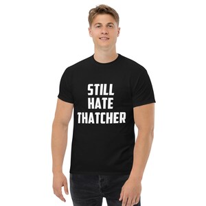 Puede incluir: Camiseta negra con el texto blanco "STILL HATE THATCHER". La camiseta es de estilo cuello redondo clásico. La persona de la imagen lleva vaqueros azul oscuro.