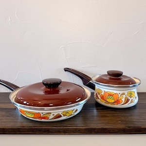 May include: Two white enameled metal pans with brown lids and handles. The exterior of each pan features a colorful floral pattern in shades of orange, yellow, and brown. The pans have a silver-toned rim and a smooth, glossy finish throughout.