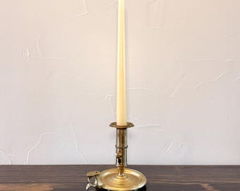 May include: A brass chamberstick candle holder with a circular base, a side handle, and a vertical stem featuring a sliding mechanism. A tall, slender, off-white taper candle is inserted into the central socket of the metallic candle holder.