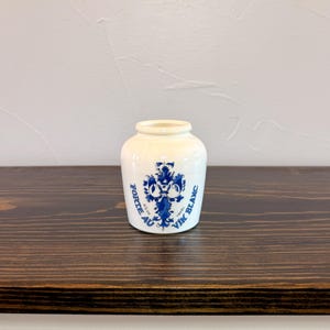 May include: A small white ceramic jar with a rounded body and a short neck. The front features a blue decorative floral emblem with the text FORTE AU VIN BLANC, 6 1/2 OZ, and 190 ML printed in blue ink around the central design.
