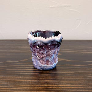 May include: A small, decorative glass vessel featuring a deep purple and white swirled pattern. The exterior surface is adorned with a complex, pressed geometric texture including starburst and diamond motifs, finished with a scalloped rim.
