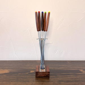 May include: A set of six metal fondue forks with dark wood handles and colorful tips in orange, red, blue, green, and yellow, held upright in a square dark wood base with a circular metal support ring.