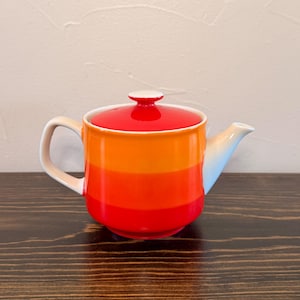 May include: A ceramic teapot featuring a horizontal gradient design that transitions from a bright orange at the top to a deep red at the base. The teapot includes a matching red lid with a small knob handle, a curved spout, and a side handle.
