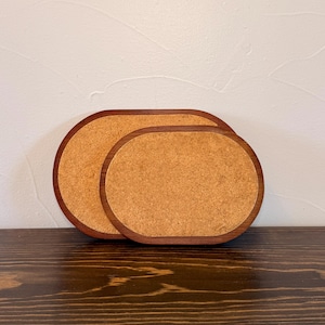 May include: Two oval-shaped coasters featuring a natural light brown cork center surrounded by a smooth, dark brown wooden frame. The coasters are layered, with the larger one positioned behind the smaller one, both displaying a simple design.
