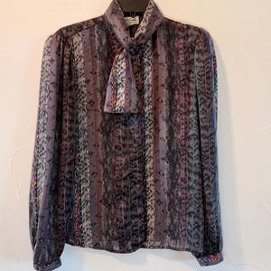 May include: A long-sleeved blouse featuring a pussy-bow neckline and a vertical striped pattern in shades of grey, black, and purple. The fabric displays a textured, abstract print with thin, colorful accents in red, blue, and green throughout the design.