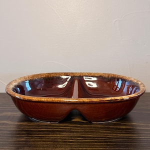 May include: A brown ceramic divided serving dish with a glossy finish. The dish has a unique shape with two separate compartments and a raised rim. The exterior is a rich brown color with a lighter brown rim.