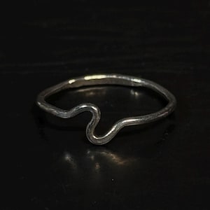 May include: A thin, silver-colored metal ring featuring a single, gentle wave-like curve along its band. The ring has a smooth, polished finish and maintains a consistent, slender thickness throughout its circular shape.