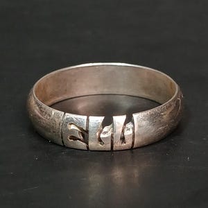 May include: A silver ring with a carved design. The ring is a band style, with a series of cut-out shapes around the circumference. The metal has a slightly aged appearance. The ring is displayed on a dark surface.