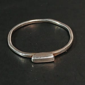 Pode incluir: Um anel de prata com uma banda fina e um detalhe retangular. O anel é apresentado sobre um fundo escuro, realçando o seu design simples e minimalista. A superfície polida do anel reflete a luz, adicionando um brilho subtil.