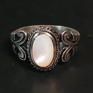 May include: A silver ring with an oval, pearlescent stone at the center. The ring features ornate, swirling designs on the sides and a braided border around the stone. The ring is set against a dark background.