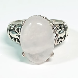 May include: A silver ring featuring an oval, pale pink gemstone. The ring's band has an ornate, openwork design. The gemstone is the focal point, set prominently in the center, creating a classic and elegant look. The ring is set against a white background.