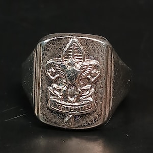 May include: A silver-toned ring with a rectangular face. The face features a detailed emblem with a fleur-de-lis and the words "BE PREPARED". The ring has a slightly worn appearance.