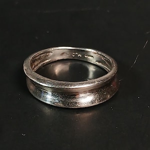 May include: A silver ring with a wide, flat band and a slightly concave inner surface. The ring has a polished finish and appears to be made of metal. The ring is displayed on a dark background.